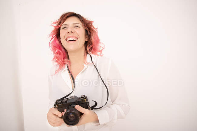 Sonríe fotógrafa con el pelo rosa sosteniendo una cámara fotográfica profesional en sus manos en la sala de luz - foto de stock