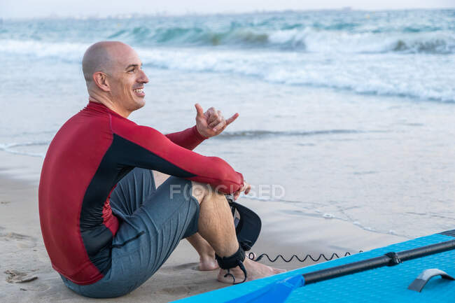 Vista lateral del pensativo surfista masculino en traje de neopreno sentado mirando hacia otro lado con tabla SUP mientras se prepara para surfear en la orilla del mar - foto de stock