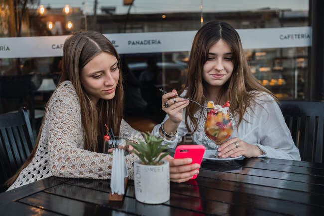 Meilleures amies avec des verres de boissons rafraîchissantes naviguant sur le téléphone portable à table dans un café urbain — Photo de stock