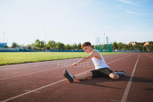 Ajuste atleta masculino en ropa deportiva inclinada hacia adelante durante el entrenamiento en pista bajo el cielo nublado a la luz del sol - foto de stock