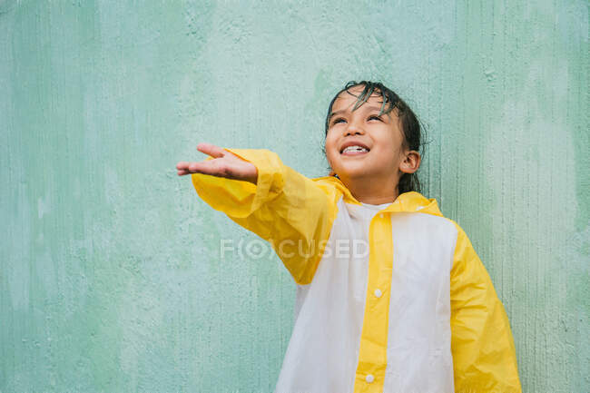 Charming ethnic child in slicker with palm up catching rain while looking up on pastel background — Stock Photo