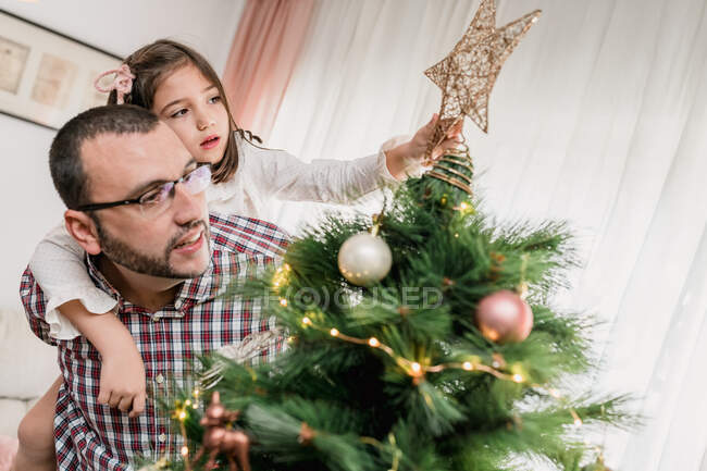 Pai dando porquinho de volta passeio para a filha decorar abeto de Natal com estrela festiva enquanto se prepara para a celebração — Fotografia de Stock