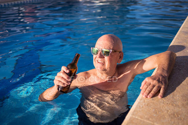 High angle of relaxed senior male in sunglasses drinking beer from glass bottle while swimming in pool — Stock Photo