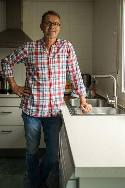 Calma hombre maduro feliz de pie mirando la cámara cerca del fregadero en la cocina mientras lava platos - foto de stock
