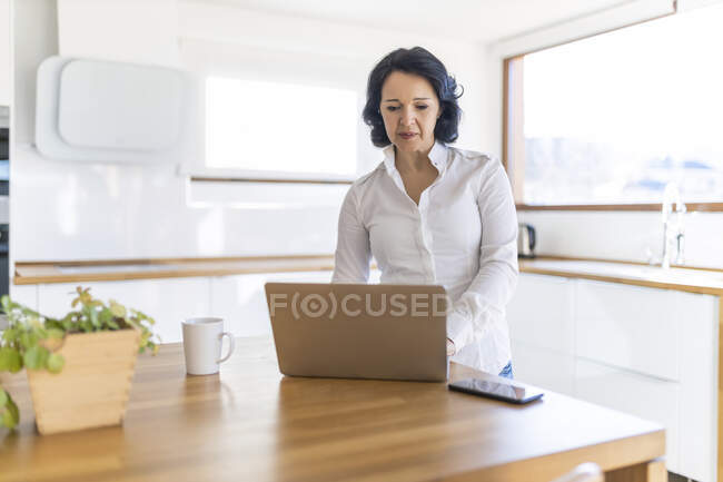 Заколдованная зрелая женщина-фрилансер, просматривающая в Интернете нетбук, работающая над новым проектом, стоя за столиком на кухне дома — стоковое фото
