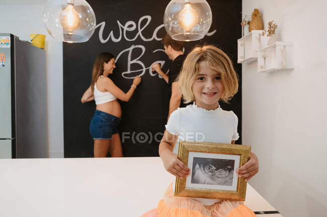 Niño encantador con futura imagen de ultrasonido hermano mirando a la cámara contra la madre embarazada y el padre escribiendo en pizarra en casa - foto de stock