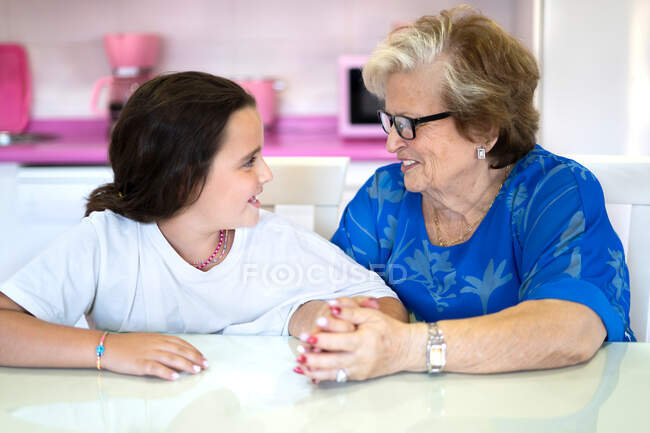 Lächelnde Großmutter in lässiger Kleidung und Brille, die am Tisch sitzt und die Hand ihrer fröhlichen Enkelin in der hellen Küche am Tag hält — Stockfoto