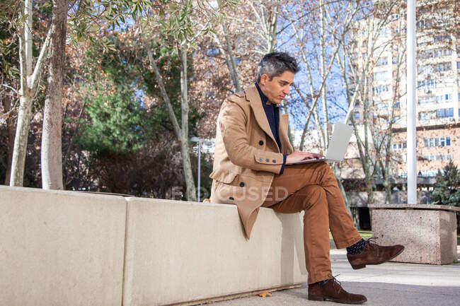 Full body of concentrated young male in formal wear sitting on bench and browsing laptop while working on project in city — Stock Photo