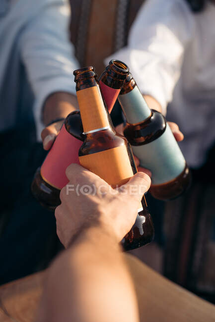 From above of crop anonymous friends clinking bottles of beer while spending weekend together in bar — Stock Photo
