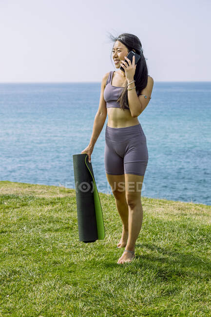 Mujer étnica joven sincera en ropa deportiva con rollo de estera de yoga hablando por teléfono celular mientras mira hacia abajo en la playa del océano - foto de stock