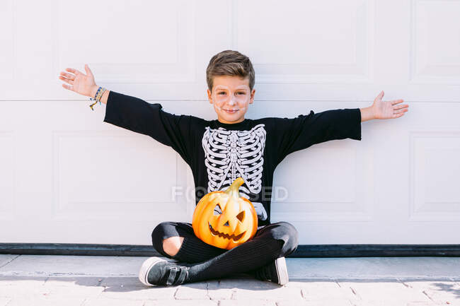 Ganzkörper aufgeregter Junge im Skelettkostüm mit Make-up und geschnitztem Halloween-Kürbis, der die Arme hebt und mit beängstigendem Gesicht schreit, während er in der Nähe der weißen Wand sitzt — Stockfoto
