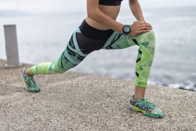 Recorte irreconocible corredor femenino estirando las piernas y haciendo ejercicio de embestida hacia adelante mientras se entrena en el paseo marítimo en verano - foto de stock