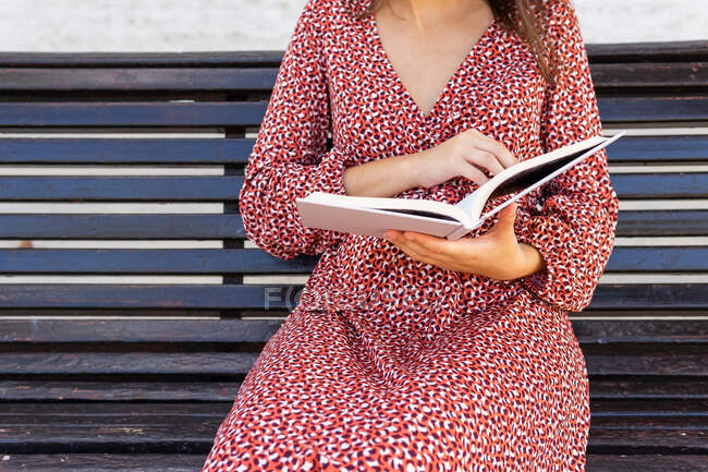 Zugeschnittene unkenntlich gemachte Frau in stilvoller Kleidung sitzt mit aufgeschlagenem Buch auf Holzbank gegen Gebäude mit Lichtwand am Tag — Stockfoto