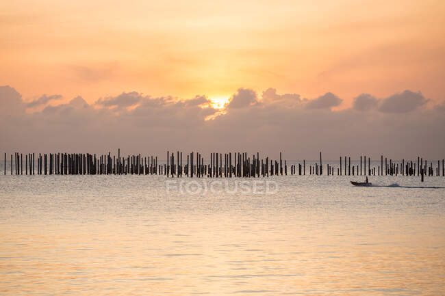 Silueta de barco flotando en ondulante mar más allá de la fila de pilares de madera bajo el cielo nublado y brillante puesta de sol en Malasia - foto de stock