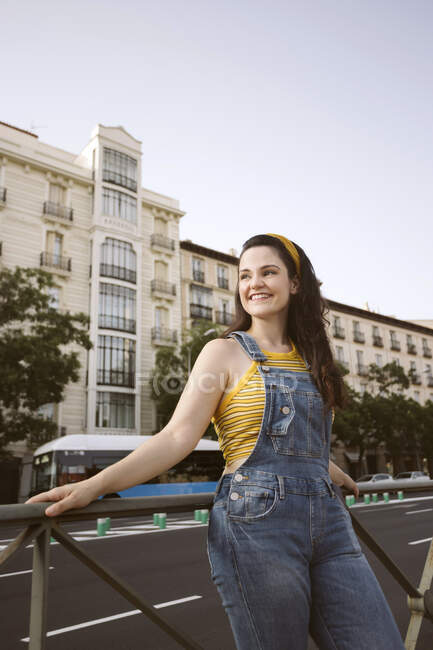 Young happy female with long brown hair wearing denim overall standing looking away with smile — Stock Photo