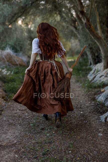 Rückansicht einer nicht wiederzuerkennenden Hexe in Kleid und mit Besenstiel, die im herbstlichen Wald entlang des Weges läuft — Stockfoto