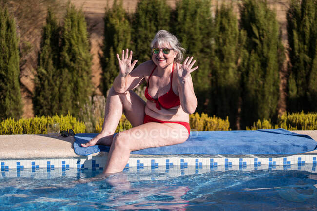 Mulher sênior alegre em biquíni sentado na toalha na beira da piscina com as pernas em água limpa e mãos onduladas — Fotografia de Stock