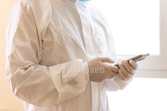 Médico masculino irreconocible recortado en equipo de protección personal que toca la pantalla en el teléfono inteligente durante la pandemia de COVID 19 en la clínica - foto de stock