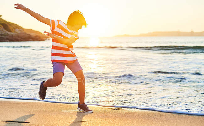 Side view full length of boy outstretching hands while running on sandy shore washed by foamy sea — Stock Photo