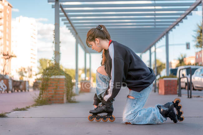 Side view of full body young female wearing black sweater and light blue jeans with slits buttoning up roller blades standing on one knee in street — Stock Photo