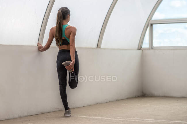 Back view of anonymous fit female athlete in sports clothes exercising with raised leg during workout — Stock Photo