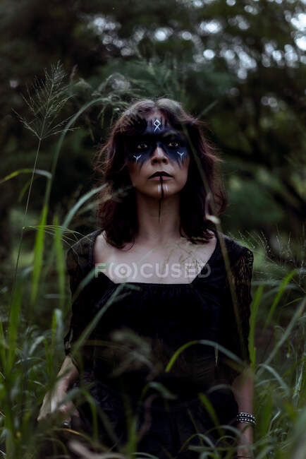 Mystic strega in abito lungo nero e con viso dipinto in piedi guardando lontano in oscuri boschi cupi — Foto stock
