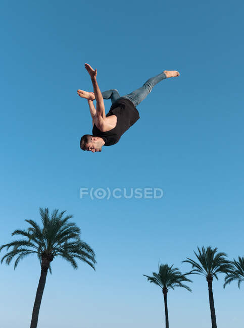Низький кут зору на активного спортсмена-чоловіка, який робить бекенд під час стрибків з пальмами під блакитним небом на сонячному світлі — стокове фото