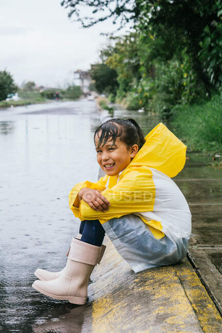 Vista laterale di felice bambino etnico in impermeabile e stivali da ginnastica seduti con le braccia incrociate sul marciapiede mentre si guarda avanti nella giornata piovosa — Foto stock