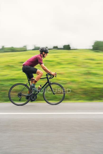 Motion blur side view of sportsman in protective helmet riding bicycle during workout on asphalt roadway against green hill and trees under light sky — Stock Photo