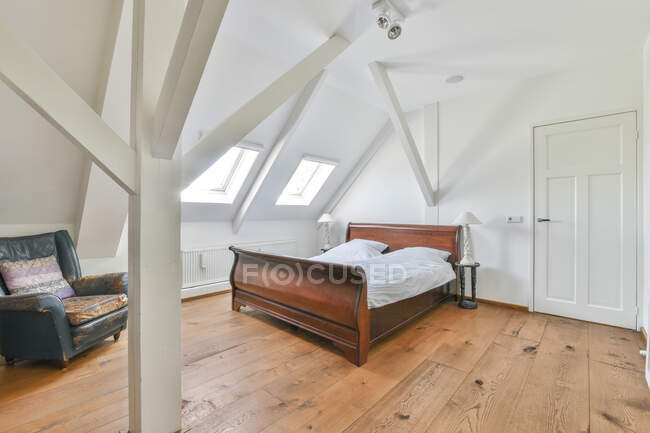 Roupa de cama na cama de madeira contra poltrona retro e janelas acima de parquet em casa durante o dia — Fotografia de Stock