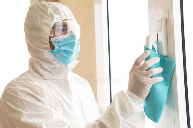 Side view of male medic in personal protective equipment cleaning handle of window with rag during COVID 19 pandemic in hospital — Stock Photo