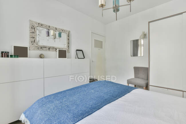 Интерьер стильной минималистской спальни с удобной кроватью, расположенной напротив белого шкафа с декоративными элементами в дневное время — стоковое фото