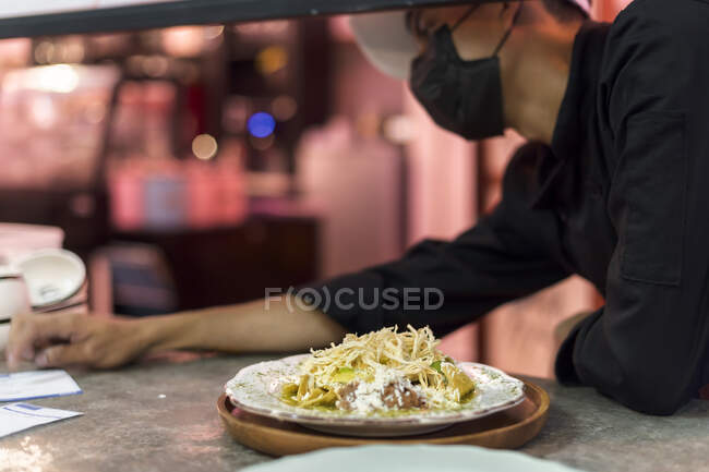 Colheita empregado masculino em máscara de tecido olhando para o balcão com prato de massa saborosa com queijo ralado e frango desfiado — Fotografia de Stock