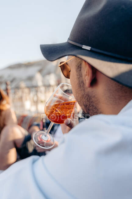 Side view of young bearded male in sunglasses and stylish hat sipping cold refreshing cocktail while relaxing on terrace — Stock Photo