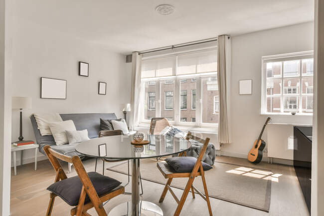 Интерьер светлой гостиной с удобным диваном и круглым стеклянным столом со стульями в дневное время — стоковое фото