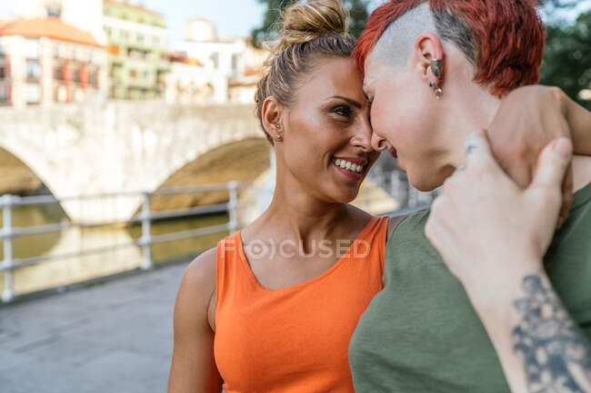 Visão traseira de mulheres homossexuais jovens com tatuagens abraçando uns aos outros tocando na testa enquanto caminham na passarela na cidade — Fotografia de Stock