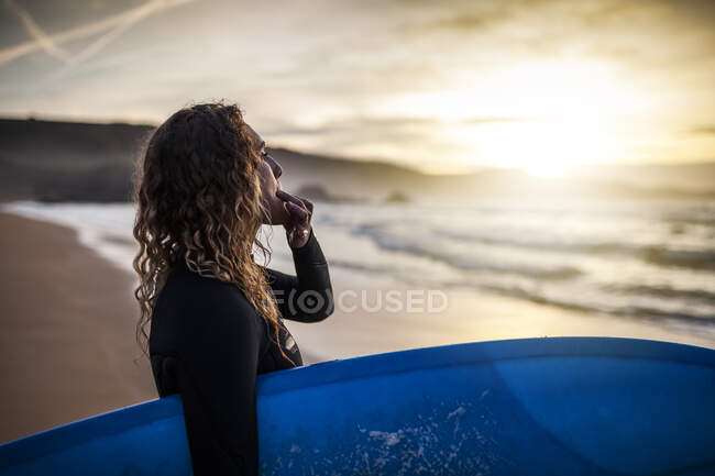 Vista lateral de la joven mujer de pie mirando hacia otro lado y silbando en la orilla con tabla de surf antes de entrar en el mar al atardecer en la playa de Asturias, España - foto de stock