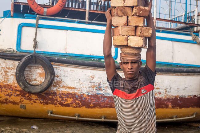 ÍNDIA, BANGLADESH - DEZEMBRO 8, 2015: Jovem etnia masculina em roupas sujas andando carregando pedras de tijolo sobre a cabeça perto do rio com barcos olhando para a câmera — Fotografia de Stock