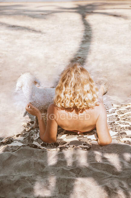 Back view of anonymous blond female traveler with dog lying on fabric on sandy shore in sunlight — Stock Photo