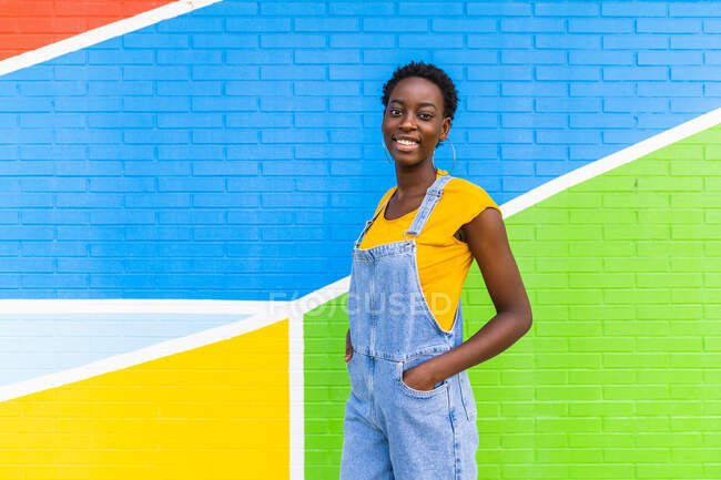 Вид на счастливую молодую афроамериканку, улыбающуюся стоя на яркой стене — стоковое фото