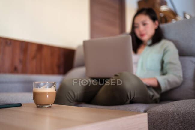 Interested young ethnic female remote employee working on netbook on sofa against coffee and smartphone at home — Stock Photo