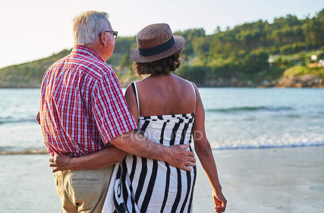 Back view of unrecognizable elderly couple in sunglasses standing on wet sandy beach and enjoying sunny day — Stock Photo