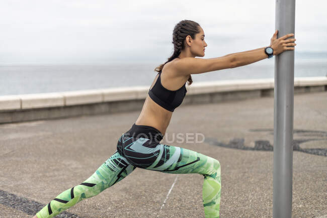 Side view of athletic female runner leaning on pillar and stretching legs while warming up on embankment during workout in summer — Stock Photo