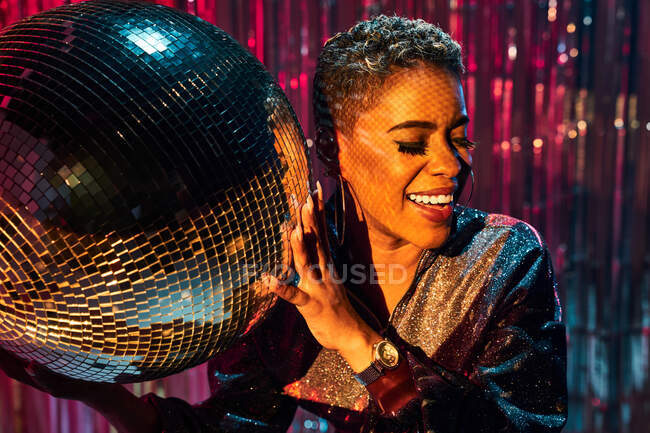 Trendy young cheerful ethnic female with eyes closed holding mirror ball in nightclub — Stock Photo