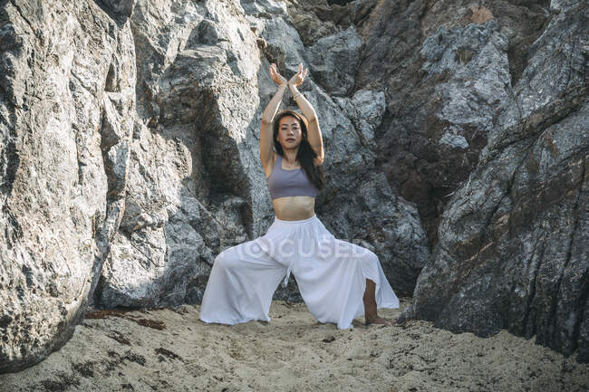 Young ethnic female in pants performing Utkata Konasana pose with raised arms while looking at camera against mount — Stock Photo