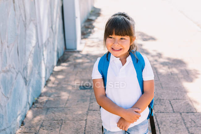 Joyeux écolier aux cheveux bruns en t-shirt blanc et au sac à dos coloré regardant loin sur une passerelle carrelée en ville — Photo de stock