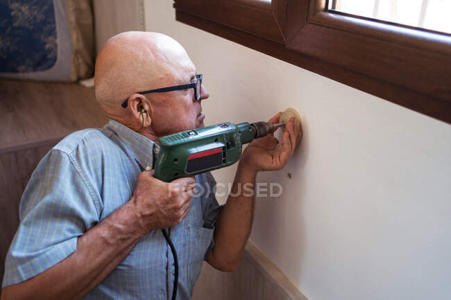Konzentrierter alter Mann in Brille mit Elektroschrauber schraubt Kunststoffteil an Hauswand — Stockfoto