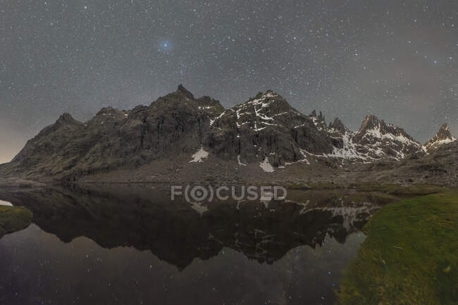 Paesaggio pittoresco di alte montagne rocciose coperte di neve che riflette in acque calme di fiume sotto cielo stellato notturno — Foto stock