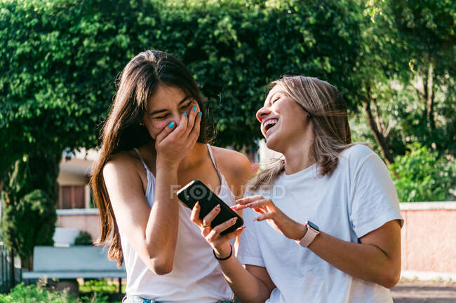 Smiling teen covering mouth while interacting with excited best female friend with cellphone in sunny town — Stock Photo