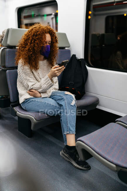 Mujer atenta con el pelo rizado en tela mascarilla navegar por Internet en el teléfono celular durante el viaje en tren - foto de stock